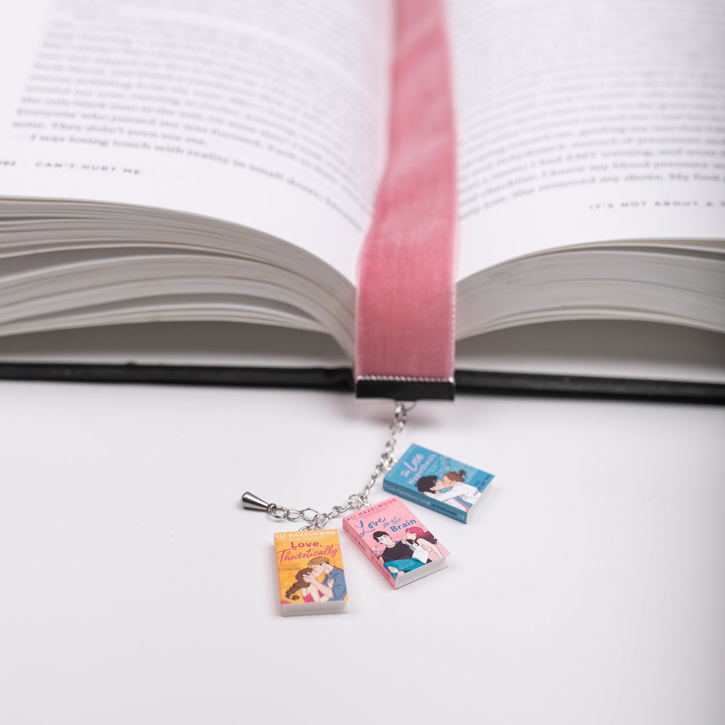 Pink, velvet ribbon bookmark with mini charms of Ali Hazelwood's books marking a page in an open book