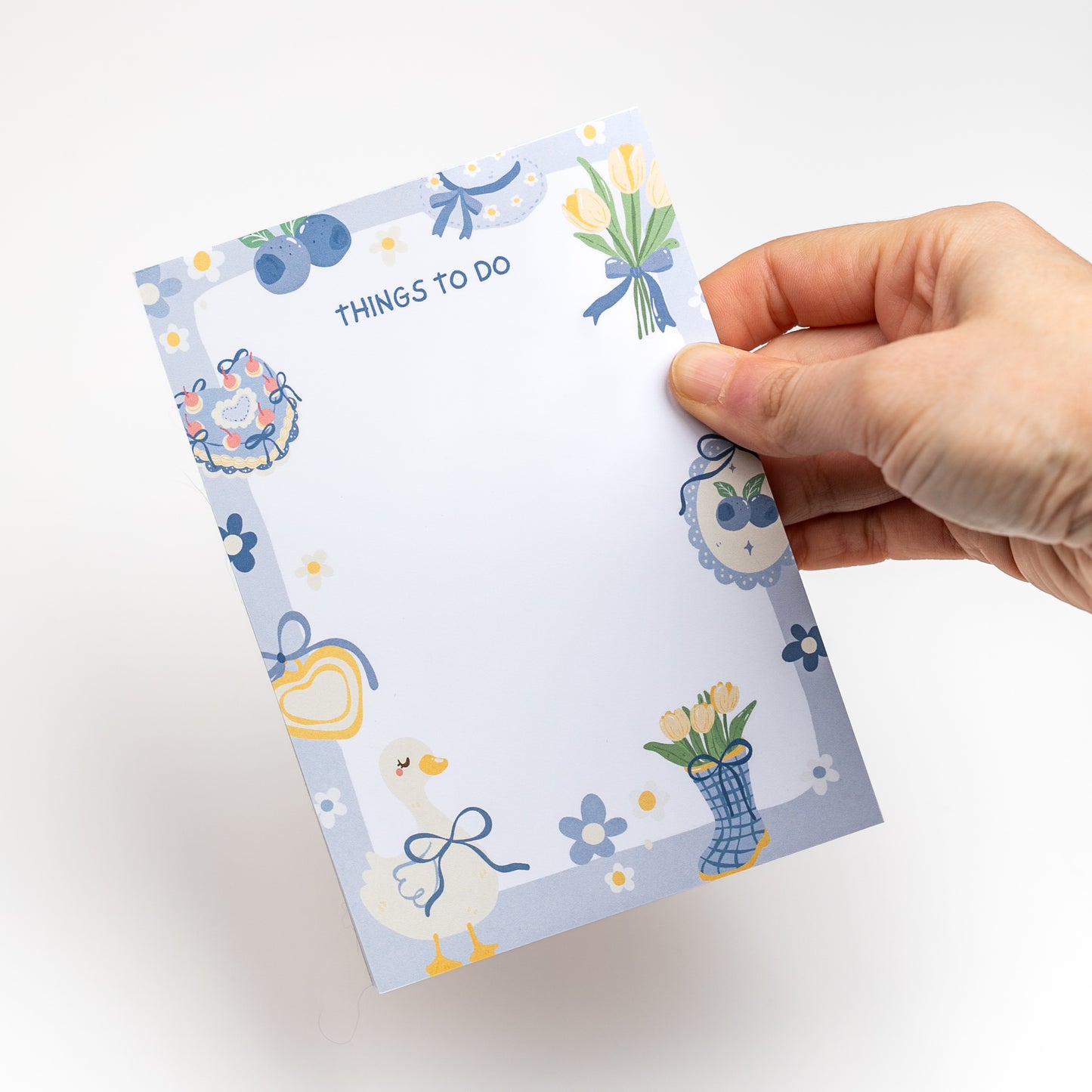 Hand holding a small notepad with decorative border and 'THINGS TO DO' text on a white background