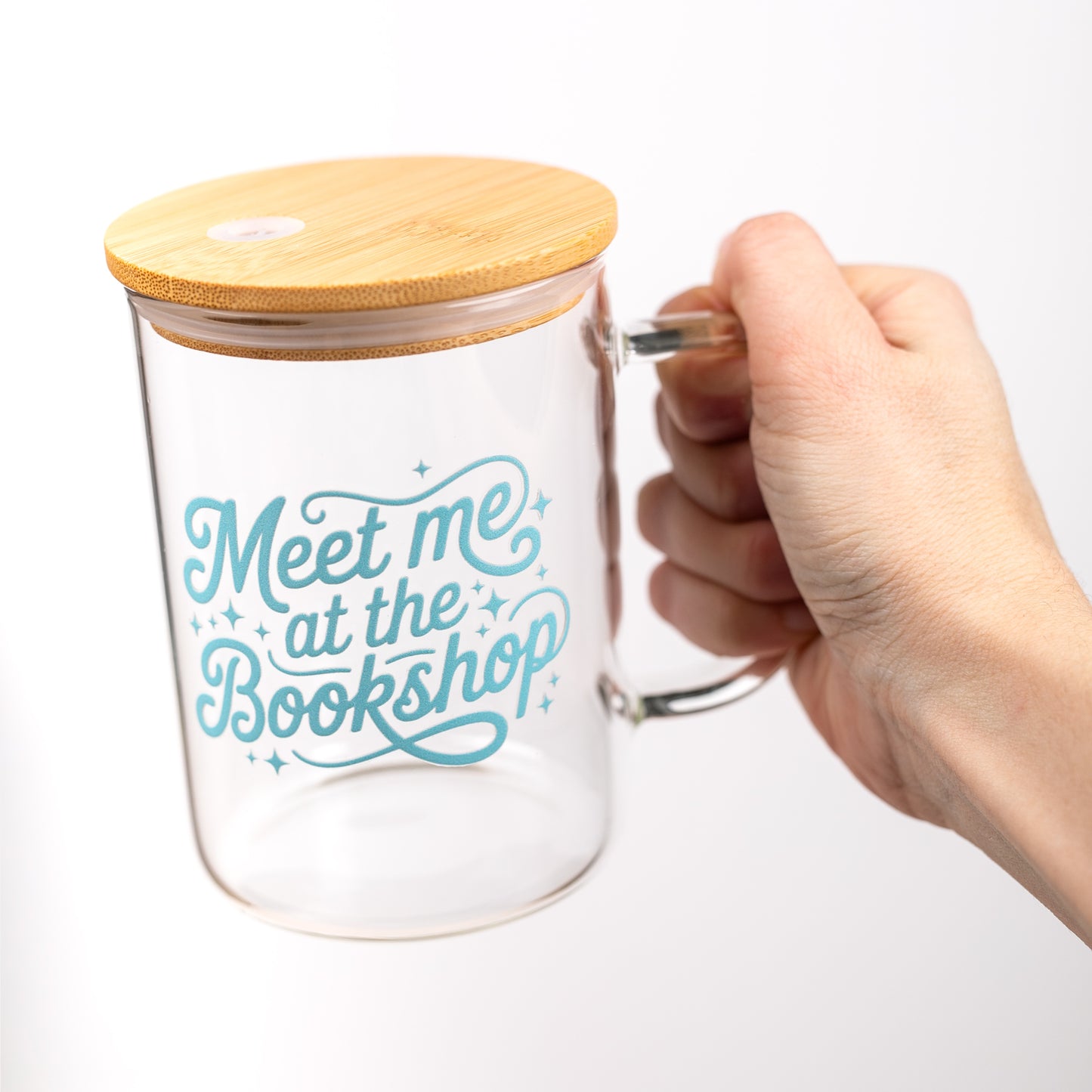 Clear glass mug with bamboo lid and 'Meet me at the Bookshop' text on a white background