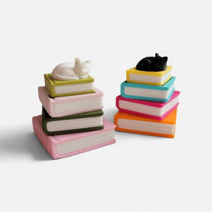 Two decorative book stacks on a white background. The left stack has pink, green, and dark green books with a white cat figurine on top.