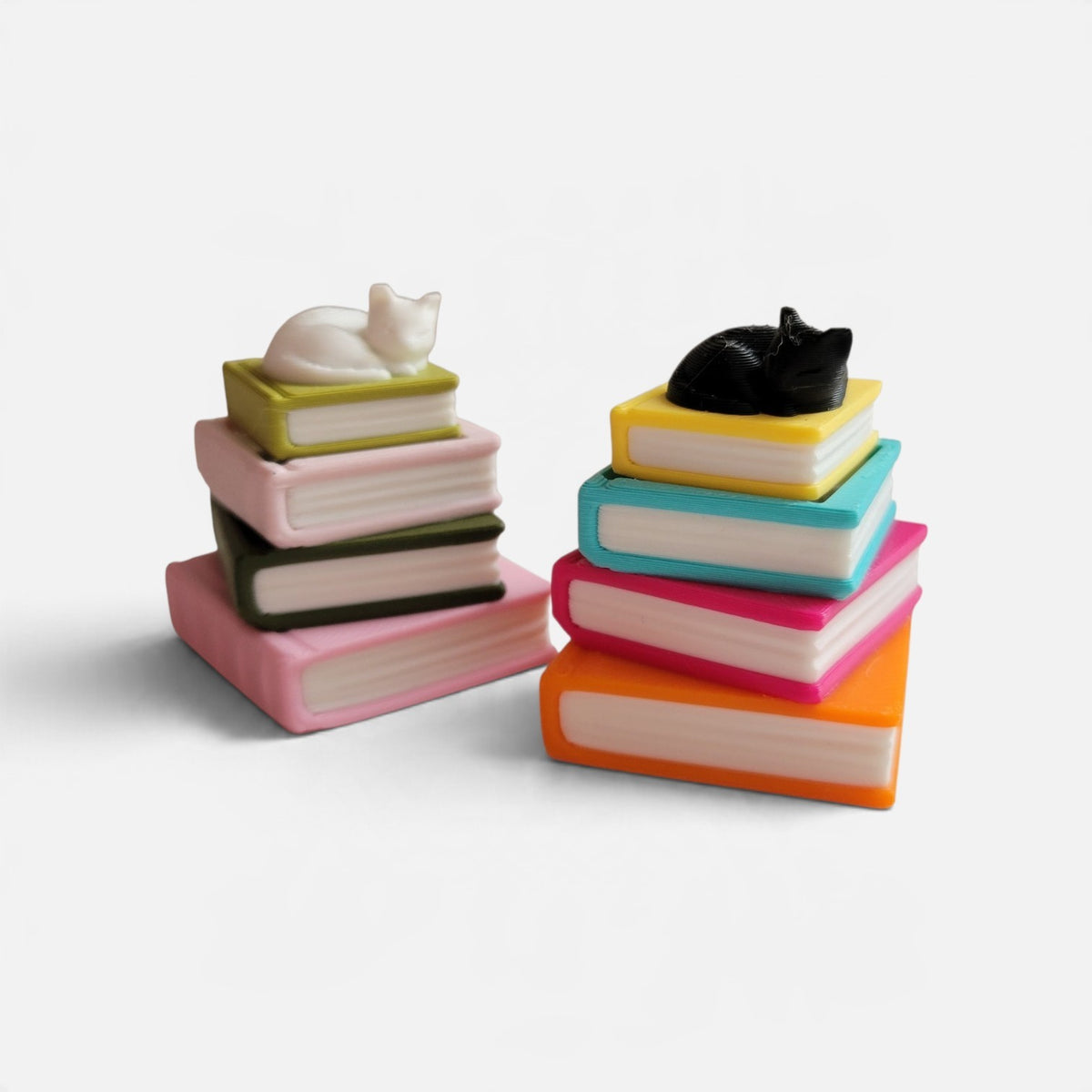 Two stacks of miniature colorful books with small cat figurines on top, photographed on a clean white background.