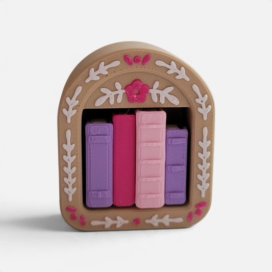 A decorative book-shaped object with a tan arched frame, white leaf motifs, pink floral accents, and three faux book spines in purple, magenta, and light pink, shown on a clean white background.