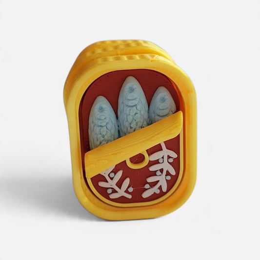A novelty jewelry piece shaped like an open yellow can of sardines, featuring three light blue sardine fillets and white Japanese kanji characters on a red background, set against a plain light gray studio backdrop.