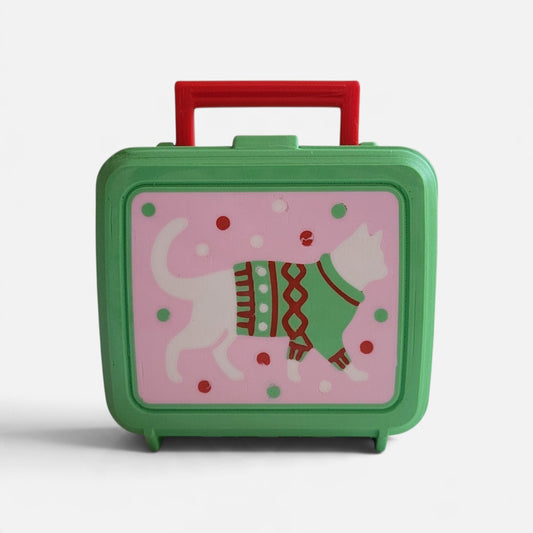 A green rolling lunchbox with a red handle, featuring a pink front panel decorated with a white cat wearing a green and red knit sweater and small red and green dots.