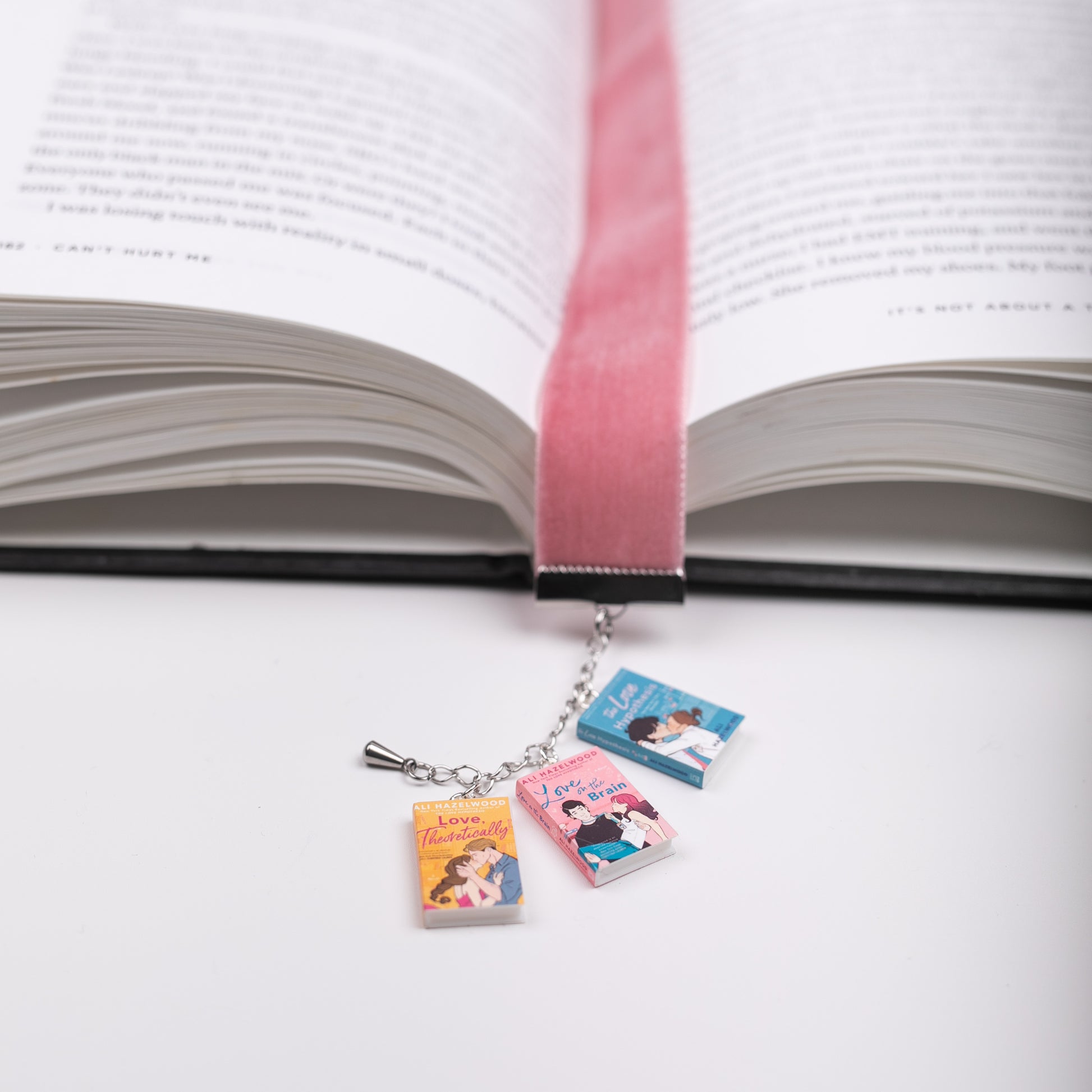 Pink, velvet ribbon bookmark with mini charms of Ali Hazelwood's books marking a page in an open book