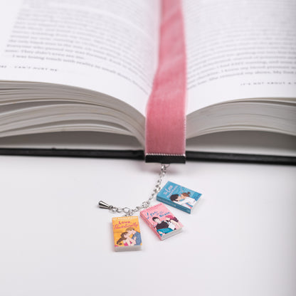 Pink, velvet ribbon bookmark with mini charms of Ali Hazelwood's books marking a page in an open book