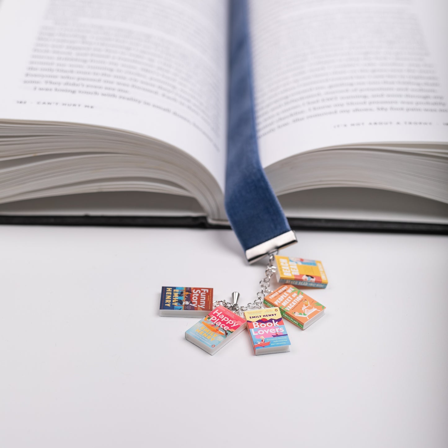 Colorful mini book charms from Emily Henry's series are featured on a blue ribbon bookmark marking the page in an open book