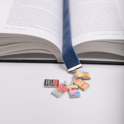 Colorful mini book charms from Emily Henry's series are featured on a blue ribbon bookmark marking the page in an open book