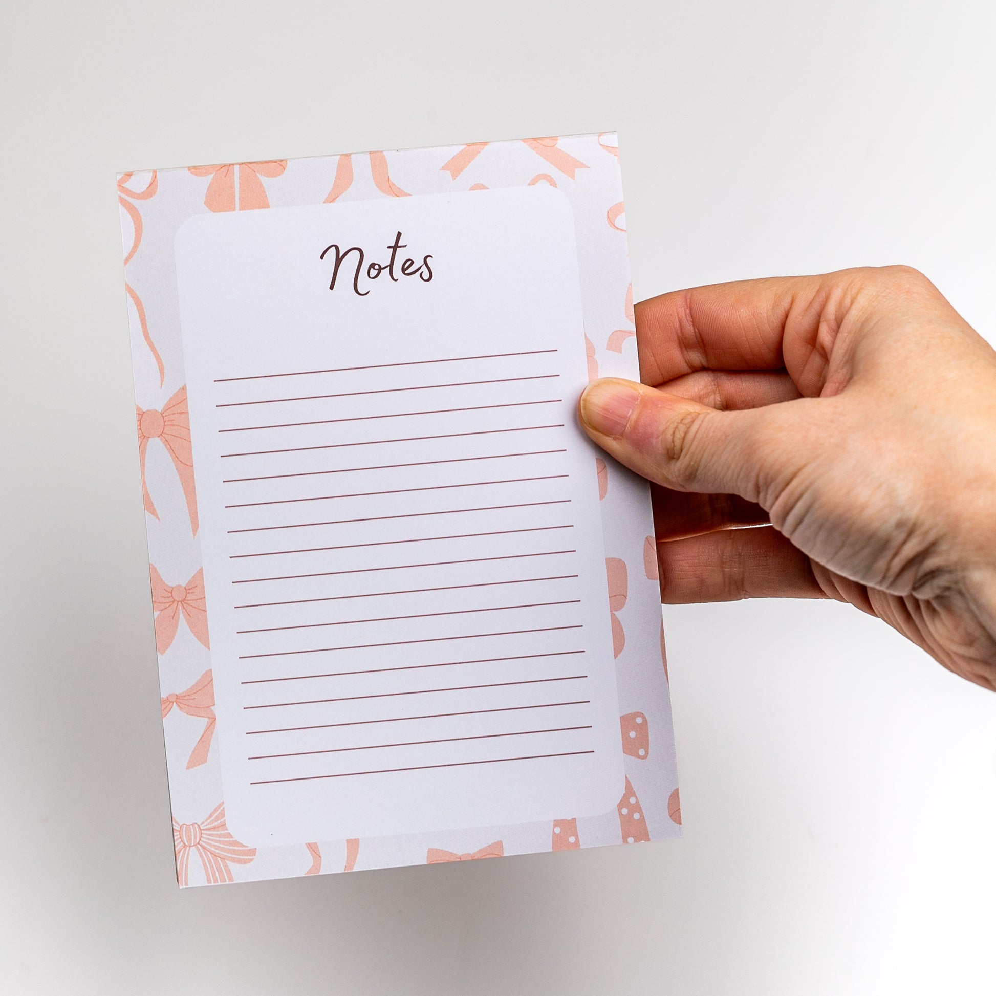 Hand holding a small notepad with 'Notes' text and pink bow design on a white background