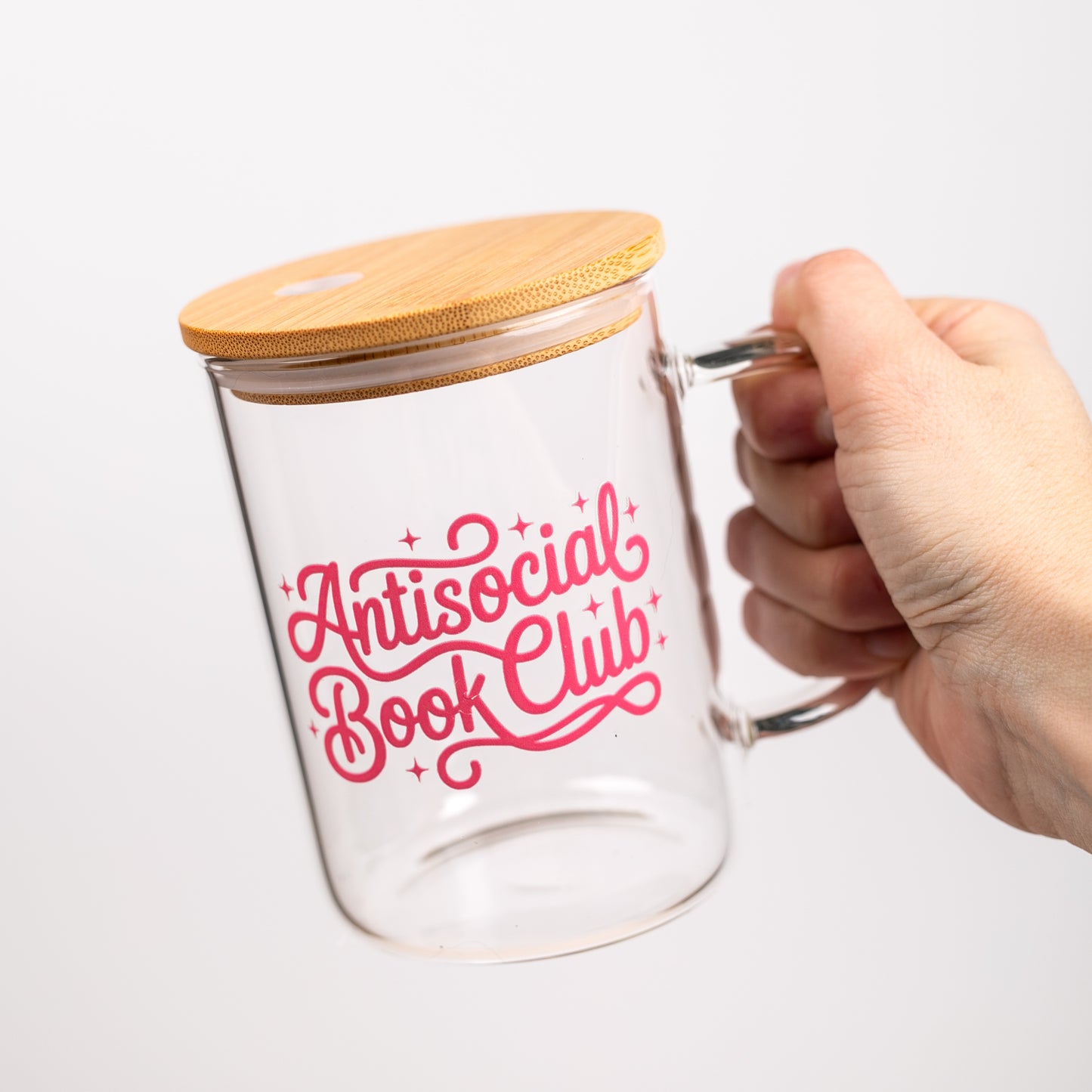 Clear glass mug with bamboo lid and 'Antisocial Book Club' text in pink on a white background