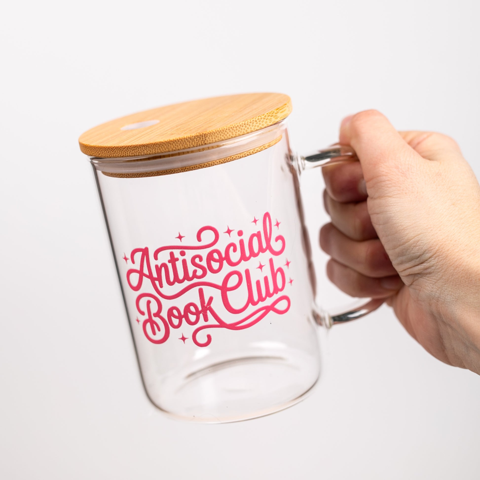 Clear glass mug with bamboo lid and 'Antisocial Book Club' text in pink on a white background