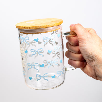 Glass mug with blue bows and hearts and a bamboo lid, held by a hand on a white background