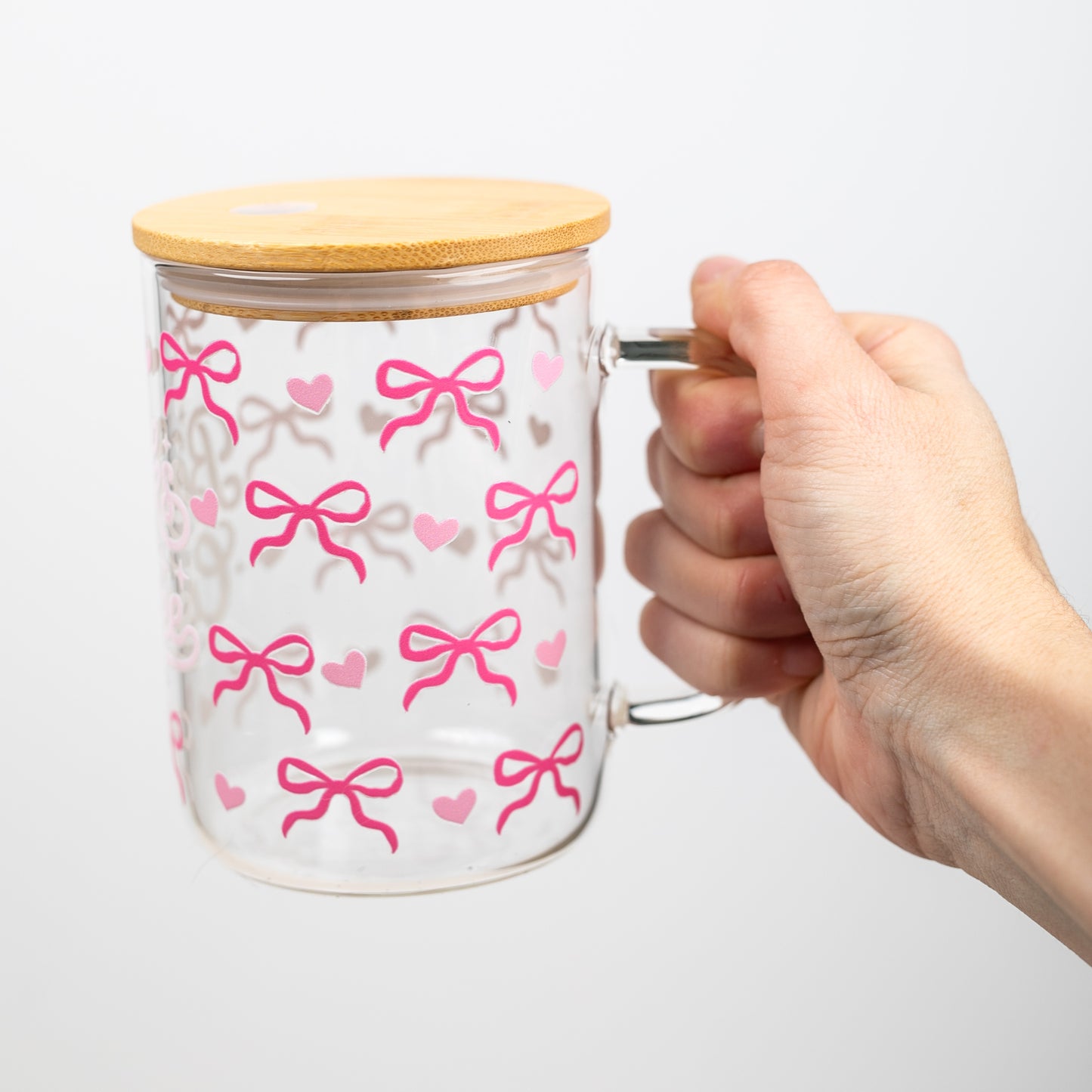Glass mug with pink bow pattern and bamboo lid held by a hand on a white background