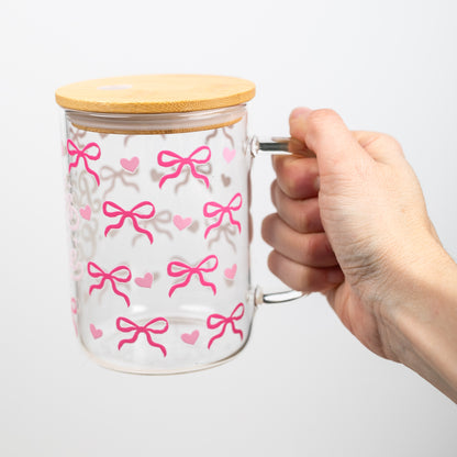 Glass mug with pink bow pattern and bamboo lid held by a hand on a white background