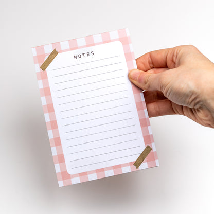 Hand holding a notepad with pink checkered border and 'Notes' text on a white background