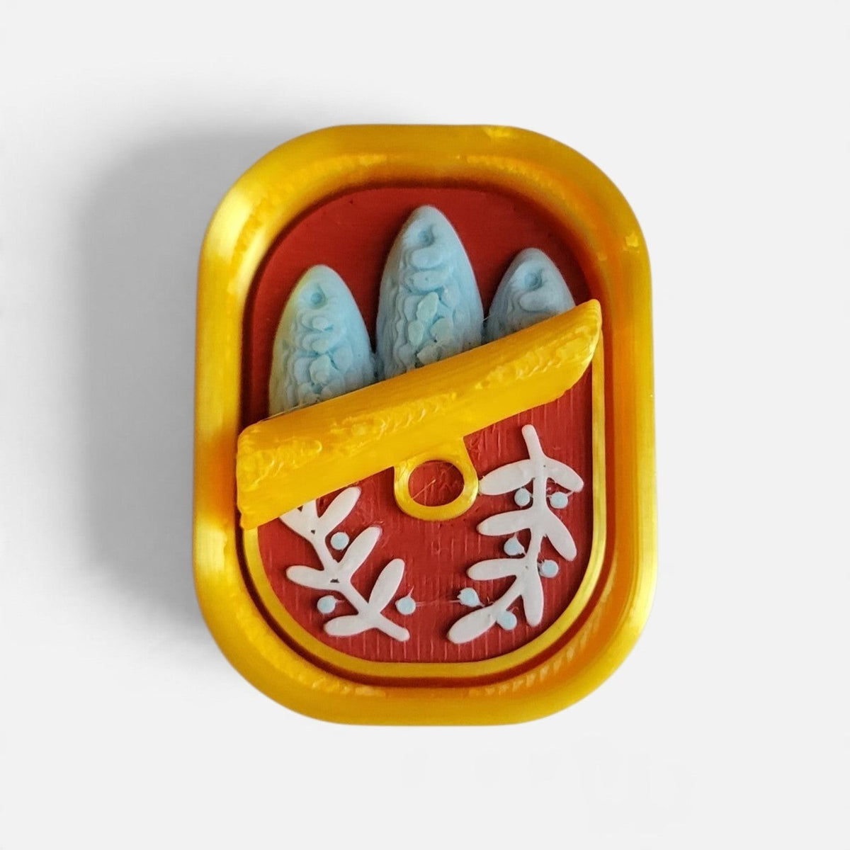 The Salty Cat Co Sardines Fidget Clicker in a yellow tin with blue fish and red background, 3D-printed sensory toy.