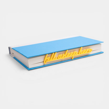 The Salty Cat Co teal Fell Asleep Here bookmark resting on a closed blue book against a white background
